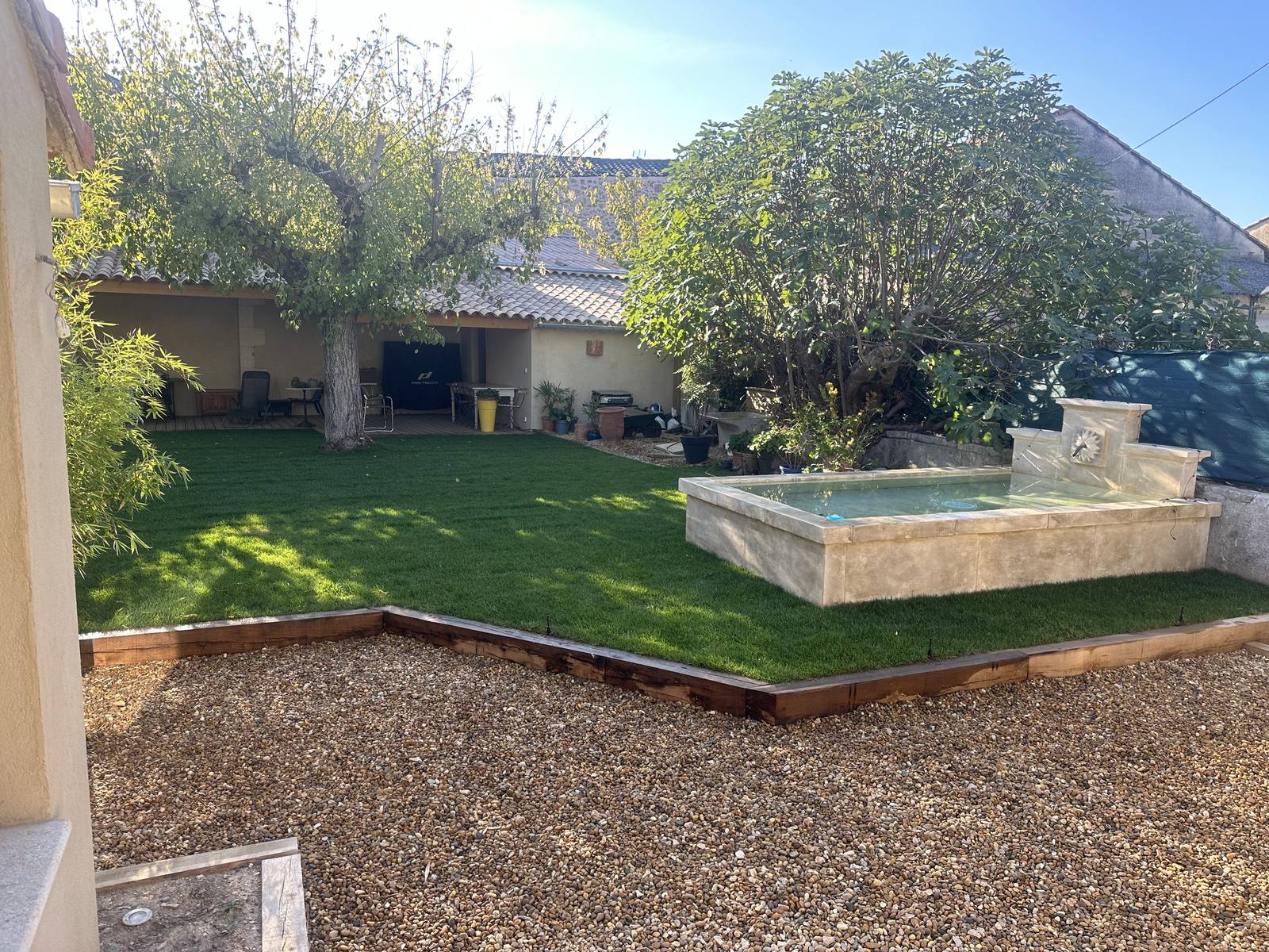 Jardin de particulier aménagé à Lauris : gazon en plaques, bordure bois, gravier et fontaine en pierre